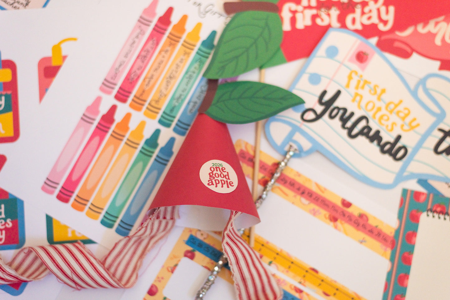 A close-up of a colorful first-day-of-school-themed party decoration set featuring a red apple shaped party hat with a white 'The Good Apple' logo, red and white striped ribbons, green paper leaves, washi tape, and sticker sheets.