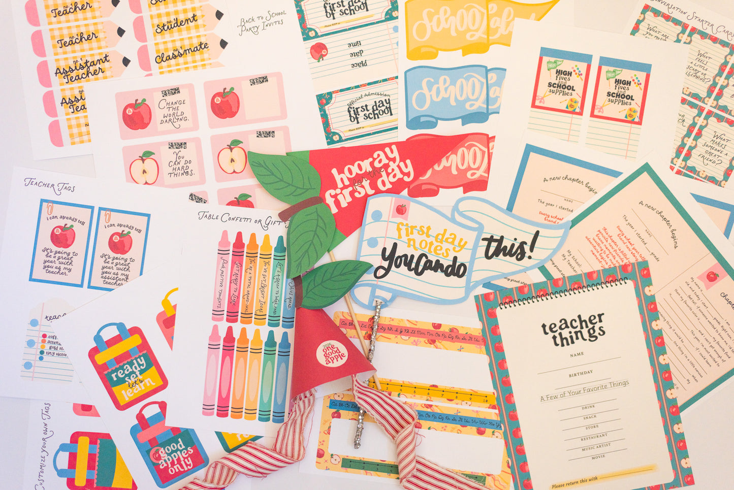 A flat-lay collection of first-day-of-school supplies and printable stationery items, including stickers, labels, a crayon box, a red 'Hooray First Day' banner, and a 'teacher things' checklist sheet, all arranged on a white background.