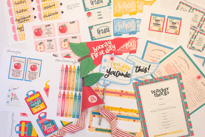 A flat-lay collection of first-day-of-school supplies and printable stationery items, including stickers, labels, a crayon box, a red 'Hooray First Day' banner, and a 'teacher things' checklist sheet, all arranged on a white background.