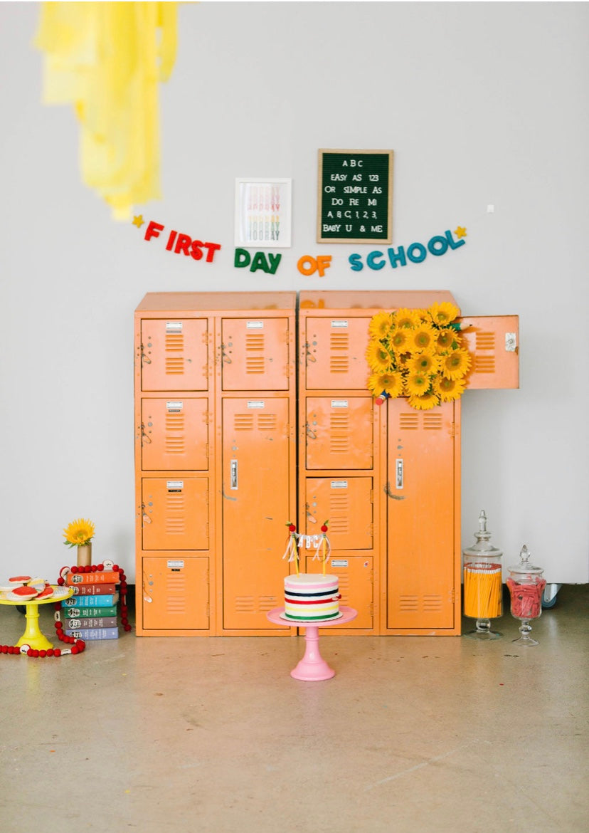 Orange lockers with 'First Day of School' sign, cake, and decorations in a classroom setting.