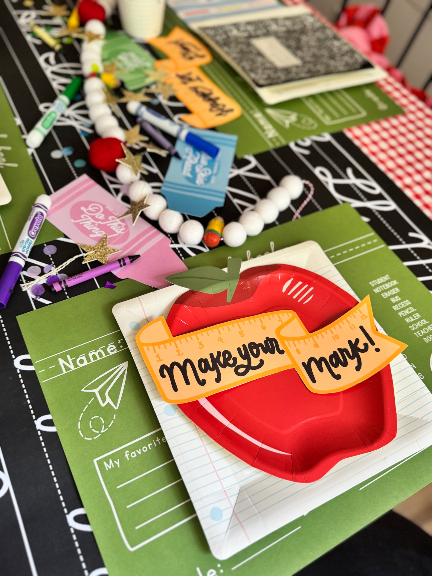 Card with red apple and 'Make your mark!' penannt flag text on a green surface with stationery items.