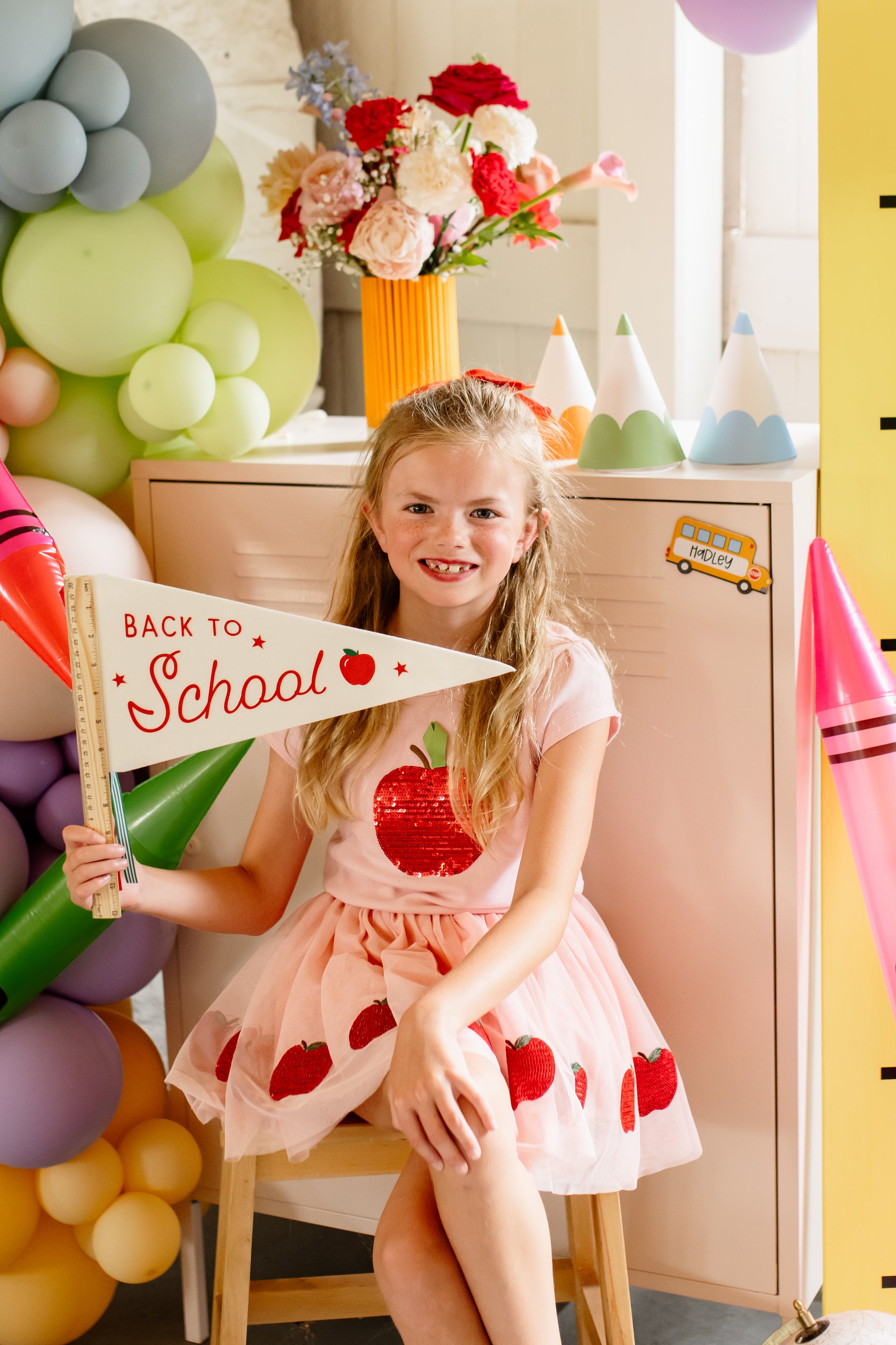 Young girl in a pink dress with red apples holding a 'Back to School' sign in a decorated room.