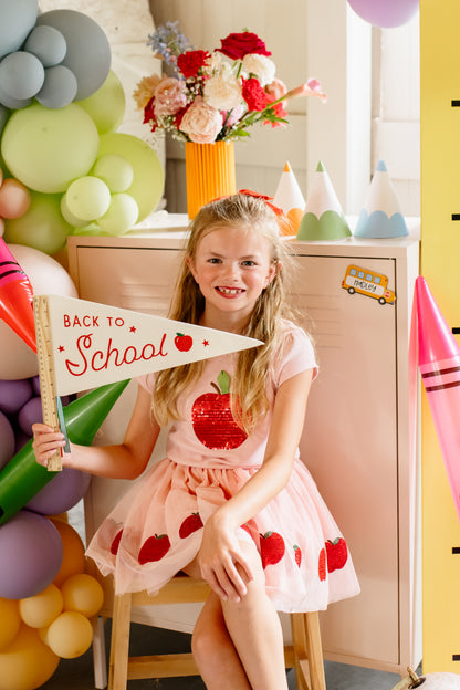 Young girl in a pink dress with red apples holding a 'Back to School' sign in a decorated room.